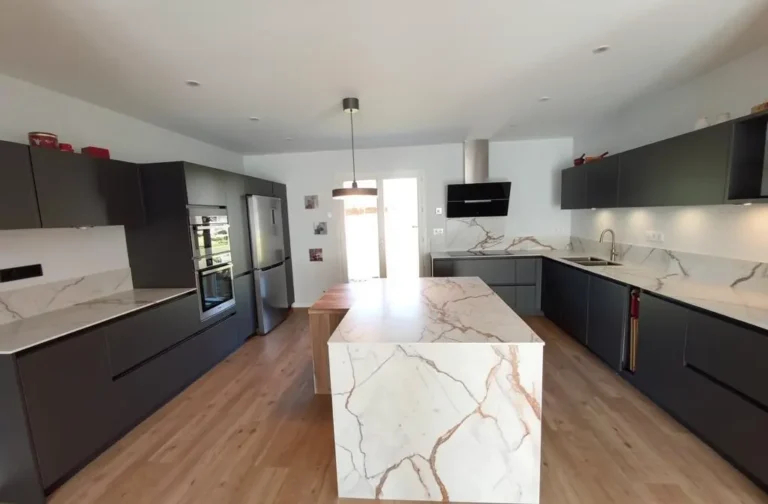 Cuisine moderne avec îlot central en marbre blanc veiné; meubles noir anthracite, plan de travail et crédence assortis, îlot central avec espace repas, four encastré et grand réfrigérateur. Aménagement cuisine avec îlot central fonctionnel et élégant.