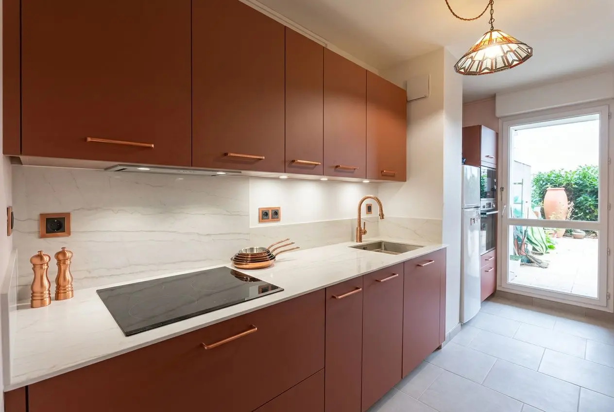 Cuisine moderne avec rangements en bois et plan de travail blanc, évier et plaque en ligne droite, porte-fenêtre laissant entrer la lumière. aménagement petite cuisine 6m2 optimisé.
