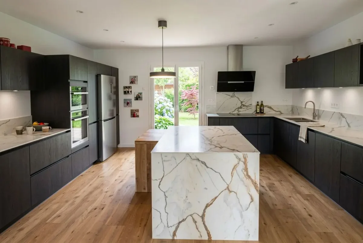 Cuisine équipée avec rangement sur mesure: îlot central en marbre, meubles noirs et plan de travail assorti, espace lumineux et accès jardin.